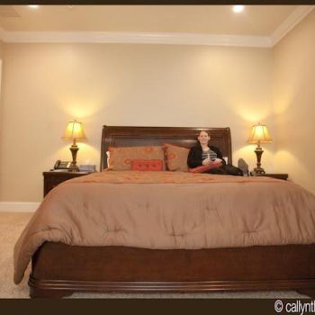 A woman sits on a large bed in a neatly arranged bedroom with two lamps on nightstands.
