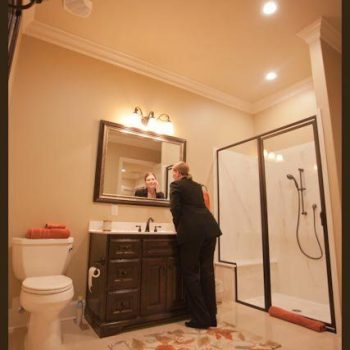 Person in a suit leaning over a bathroom sink, looking into a mirror, with a shower and toilet visible nearby.