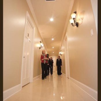 Four people stand in a well-lit hallway with beige walls and white doors, two wall sconces visible on the right.