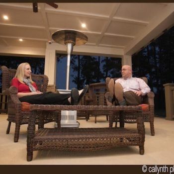 Two people sit and relax in wicker chairs on a covered patio in the evening, with feet resting on a glass-top table.
