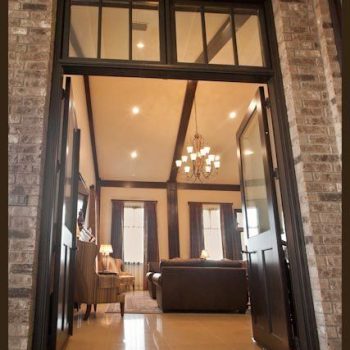 Open double doors lead from a brick exterior into a warmly lit living room with sofas and a chandelier.