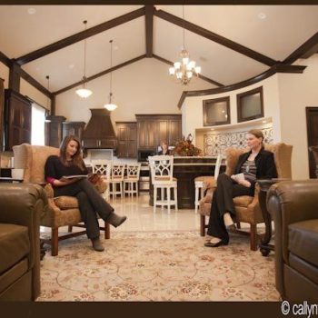 Two women sit in armchairs having a conversation in a spacious, modern living room with an open kitchen.