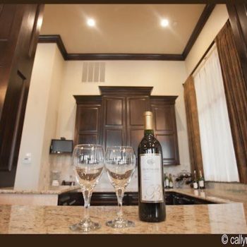 A bottle of wine and two empty wine glasses on a granite kitchen counter with dark wood cabinets in the background.