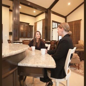 Two women sit at a kitchen island with coffee mugs, having a conversation in a modern home interior.