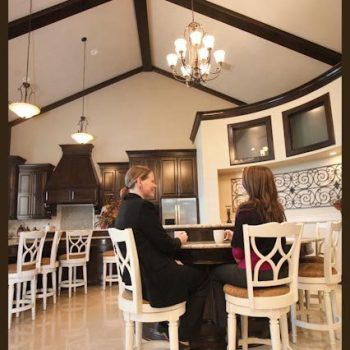 Two people sit at a round table in a spacious, modern kitchen with high ceilings and elegant lighting.