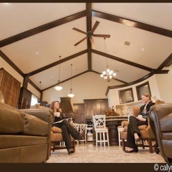 Two women sit and talk in a spacious living room with vaulted ceilings and an open kitchen in the background.