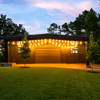 Modern metal building with string lights, large garage door, and manicured lawn surrounded by trees at dusk.