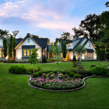 A modern suburban house with large windows, manicured lawn, flower beds, and surrounding trees at sunset.