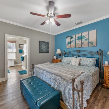 Bedroom with blue accent wall, large bed, wooden nightstands, TV, ceiling fan, and view into a connected bathroom.