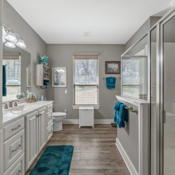 Modern bathroom with double sinks, a glass shower, wooden floors, gray walls, and teal accents.