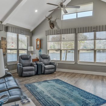 A living room with gray furniture, large windows, wood floors, a rug, and a mounted deer head above the windows.