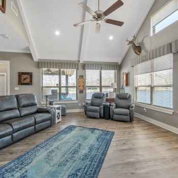 Spacious living room with gray leather seating, blue rug, wood floors, large windows, and a ceiling fan.
