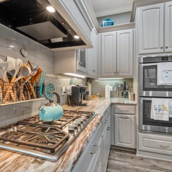 A modern kitchen with a gas stove, blue kettle, double ovens, stainless fridge, and gray cabinets with utensils on display.