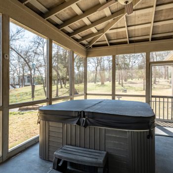 A covered screened porch with a closed hot tub, ceiling fan, and views of a grassy backyard with trees.