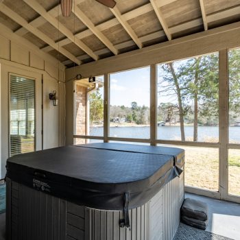 Screened porch with a covered hot tub, overlooking a lake and trees outside on a sunny day.