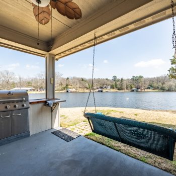 Covered patio with a grill, hanging swing, utensils, and a view of a lake with trees in the background.