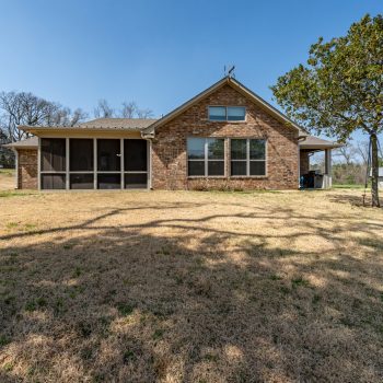 Brick house with large windows and screened porch, set on a grassy yard with trees and a clear blue sky.
