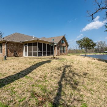 Single-story brick house with screened porch, situated on a grassy yard beside a pond under a clear blue sky.