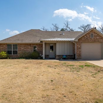 Single-story brick house with attached garage, front porch, and dry lawn under a clear sky.