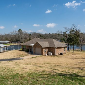 Single-story brick house with a driveway and boat dock, situated near a lake with trees and blue sky in the background.